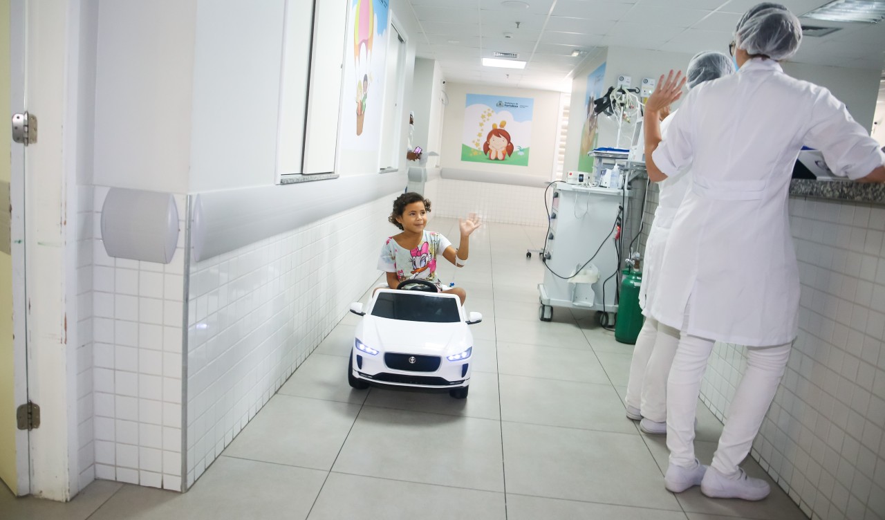 uma menina, no carrinho motorizado, cumprimenta enfermeiras num corredor do IJF